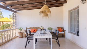 Un porche techado con una mesa y sillas blancas, una lámpara colgante de fibras tejidas y plantas en macetas.