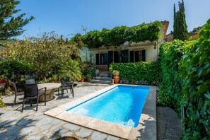 A swimming pool with stone tiles, surrounded by greenery and a building with a stone staircase.