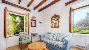 Una habitación con un sofá azul, un sillón de mimbre, una mesita, un ventanal con contraventanas abiertas, un espejo y un radiador.