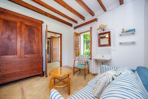 A room with a large wooden wardrobe, a wicker table, a striped sofa, a chair, and a window with a view of greenery.