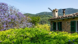 Un edificio de piedra con contraventanas verdes, rodeado de abundante follaje y flores púrpuras.