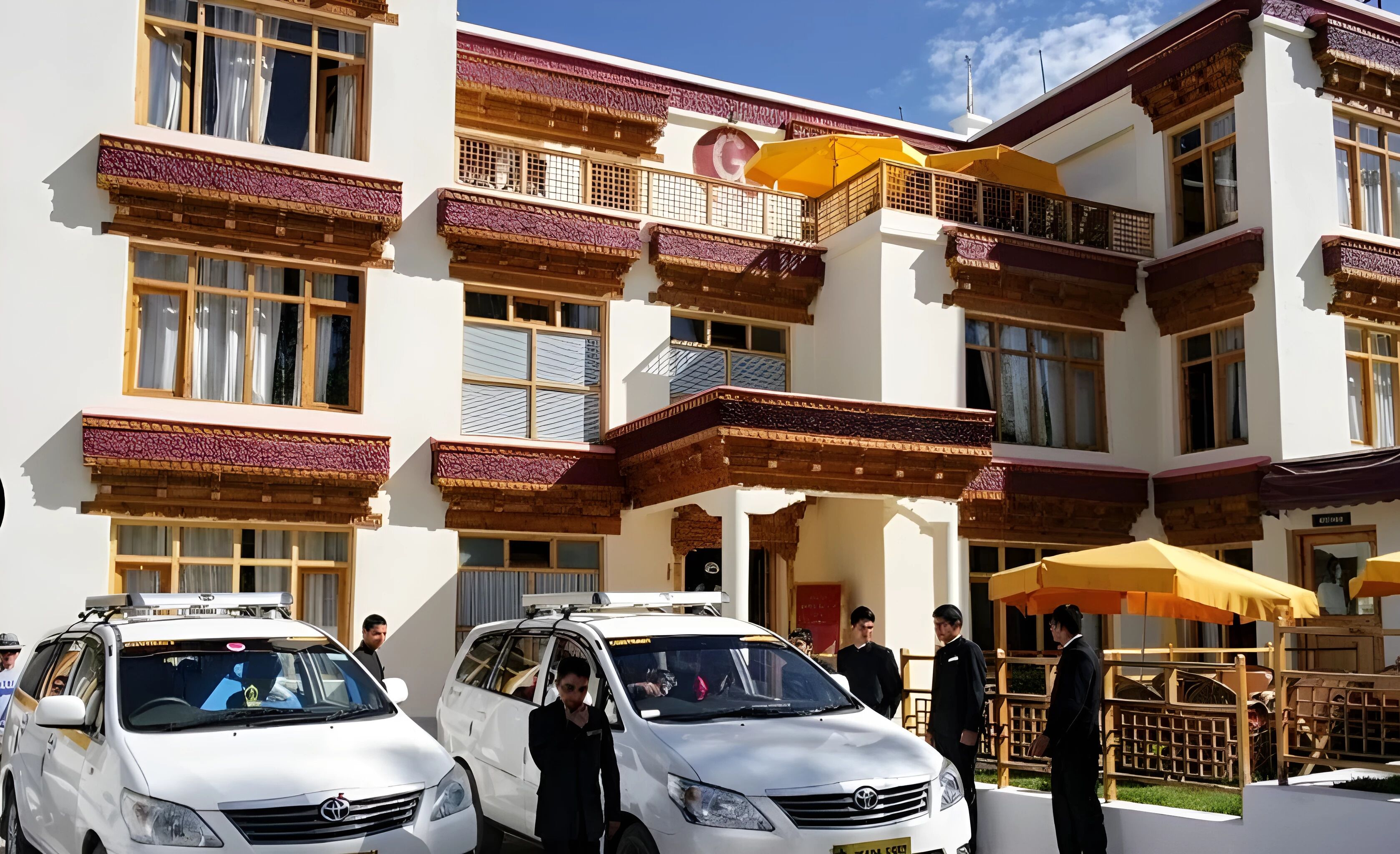A building with traditional architecture, featuring balconies with intricate designs, a central entrance with a canopy, and parked white vehicles.