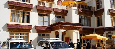 A building with traditional architecture, featuring balconies with intricate designs, a central entrance with a canopy, and parked white vehicles.
