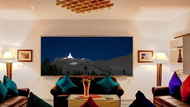 A living room with a large flat-screen TV on the wall, two leather sofas with teal and purple cushions, a coffee table with a vase, and two floor lamps. - Gomang Boutique Hotel  (Leh)