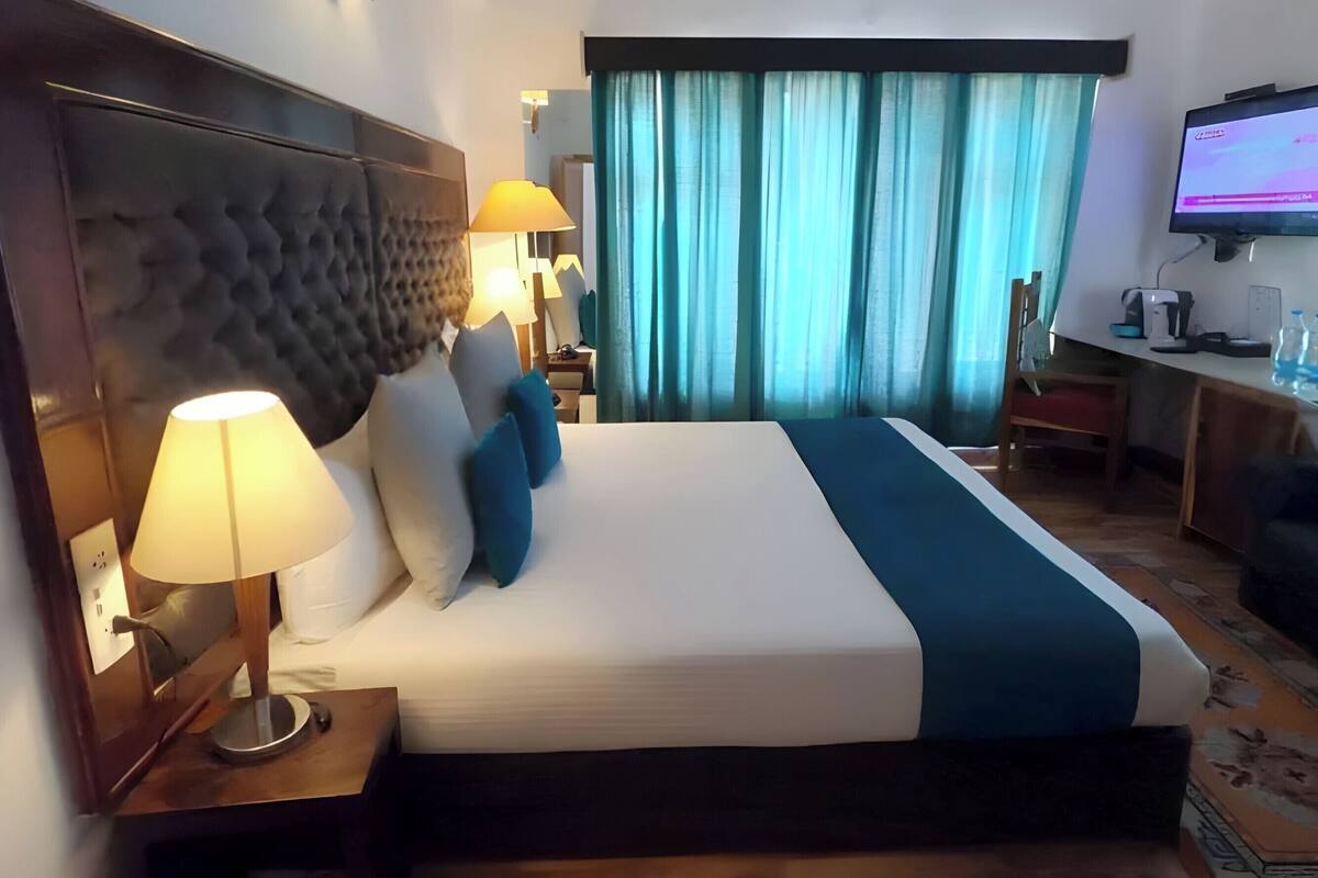 Ein Hotelzimmer mit einem großen Bett, einem Kopfteil, einem Schreibtisch mit Computer, einem Fernseher und einem Fenster mit Vorhängen.