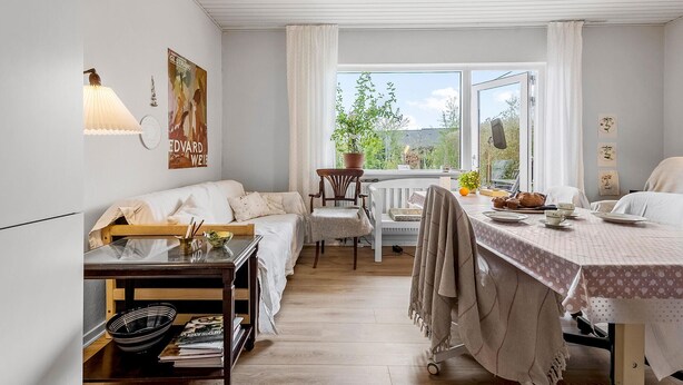 A cozy room with a wooden floor, a sofa, a dining table with a polka dot tablecloth, and a window with a view of greenery.