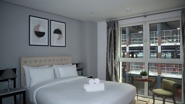 A hotel room with a large bed, a small table with a plant, and a view of a building through a large window.