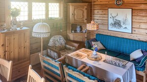 A cozy cabin interior with a wooden dining table, chairs, a sofa, and a window with decorative glass panes.