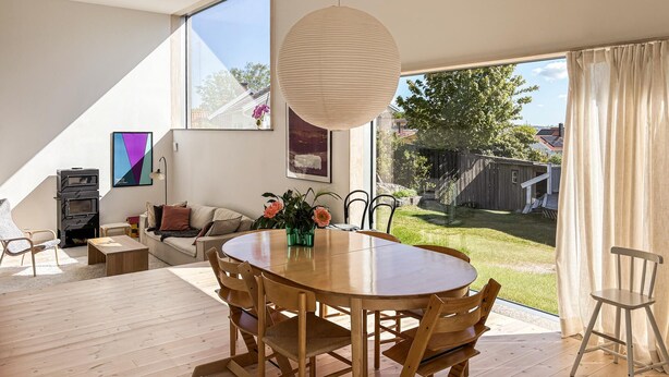A modern dining area with a wooden table and chairs, a hanging light fixture, and a view of a backyard with a wooden fence and greenery. - Stunning home in Brännö (Brännö)