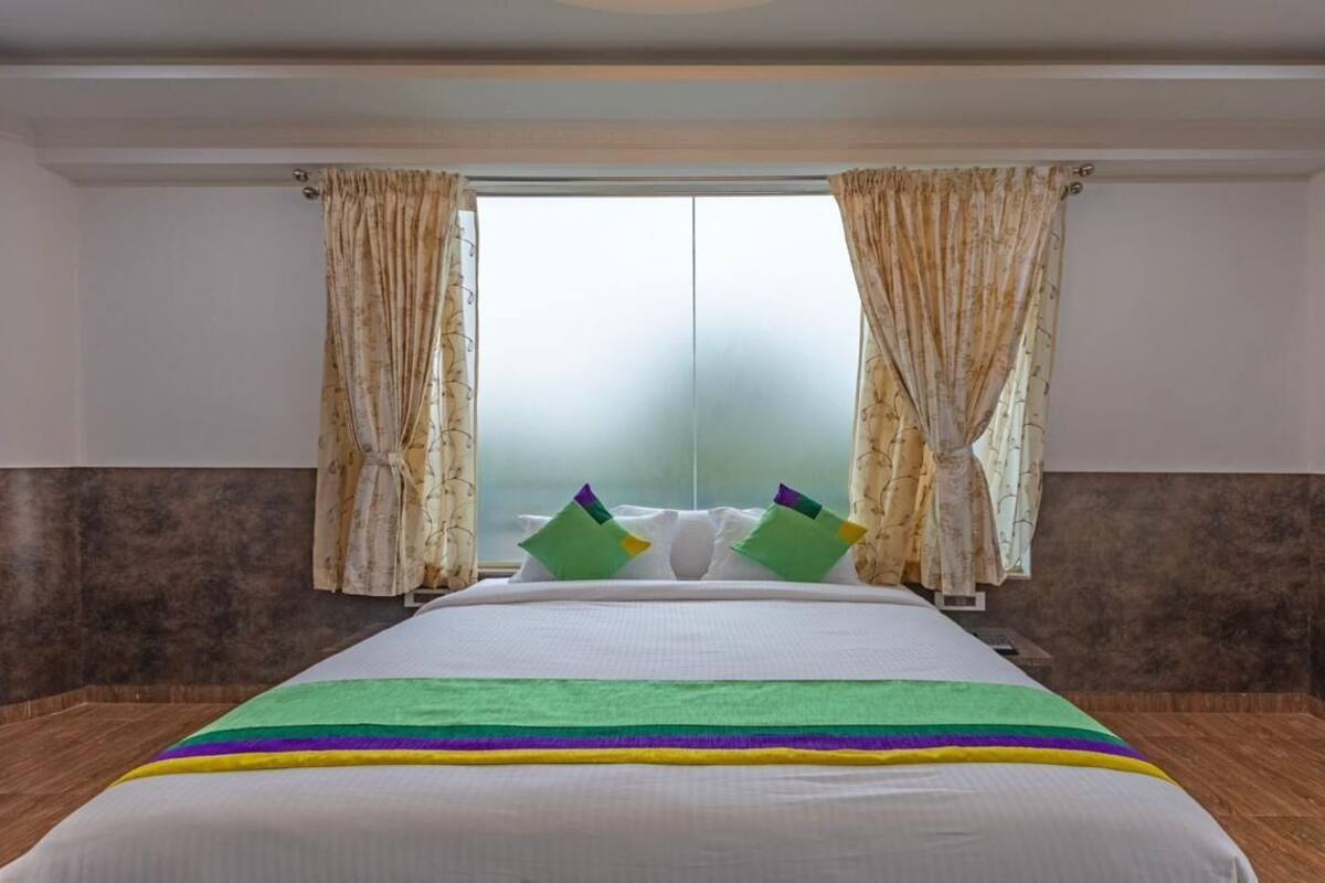 Een hotelkamer met een groot bed, beige gordijnen en uitzicht door een matig doorschijnend raam.