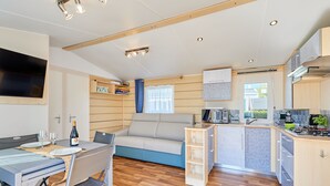 A compact kitchenette with a stove, microwave, and sink. There is a dining area with a table and chairs, and a sofa with a window. - Super Octalia (camping 5*) by Interhome (Les Pieux)