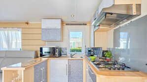 A compact kitchen with a microwave, toaster, and stove, wooden cabinets, and a window overlooking a residential area. - Super Octalia (camping 5*) by Interhome (Les Pieux)