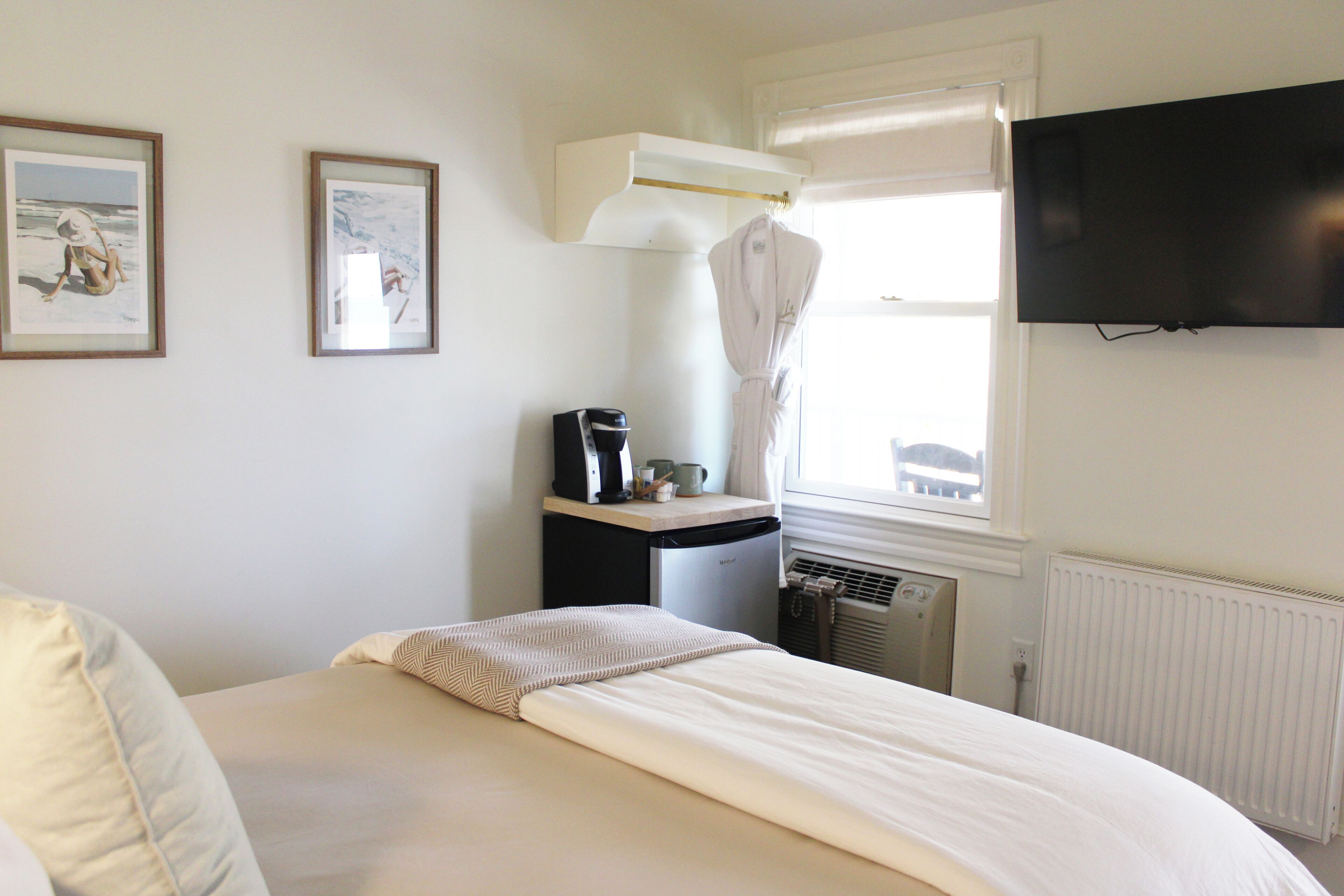 A bedroom with a bed, a desk with a coffee maker, a radiator, and a TV on the wall.