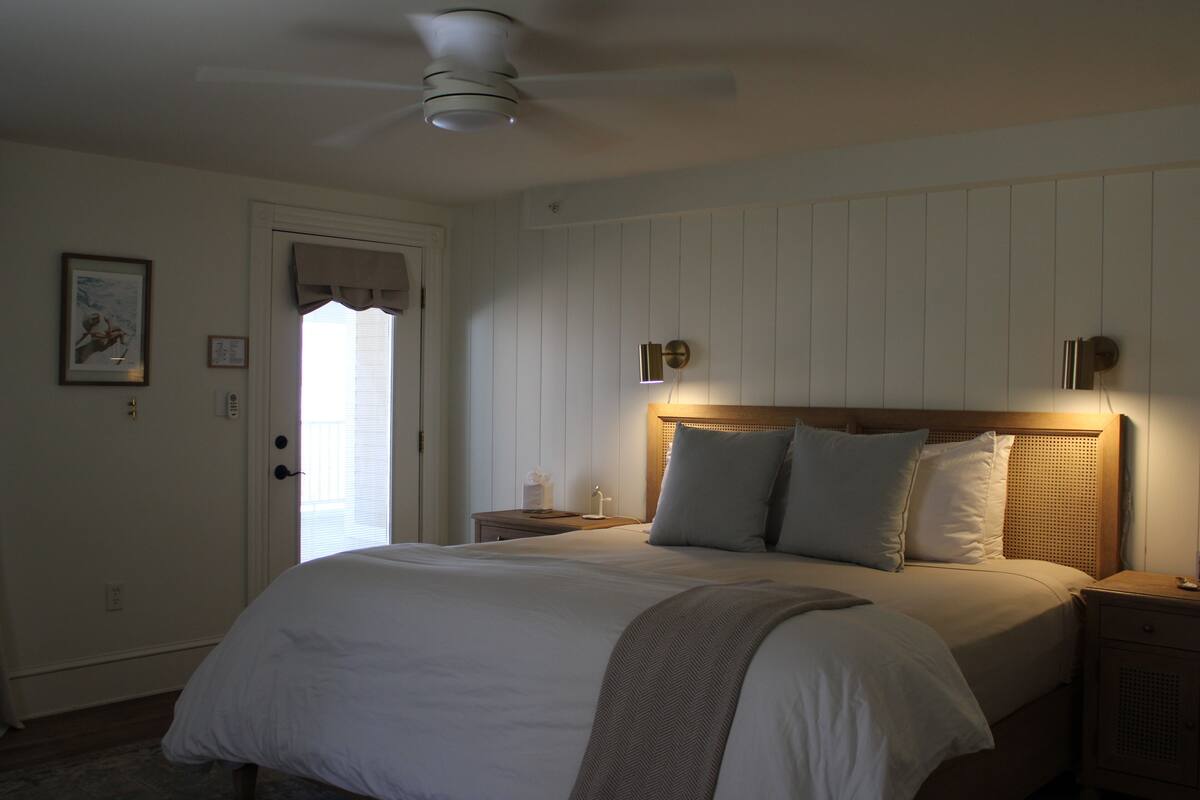 A bedroom with a bed, a nightstand, a wall-mounted light, a ceiling fan, and a door with a window.