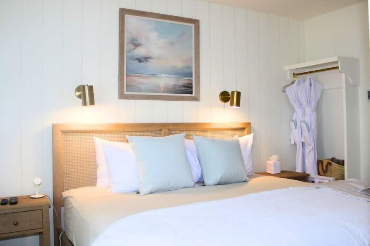 A hotel room with a bed, bedside table, lamp, robe, and a painting on the wall.