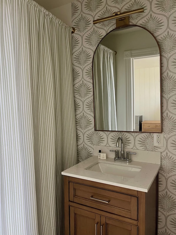 A bathroom with a striped curtain, a mirror, a sink, and wooden cabinets. - The Trident Inn (Ogunquit)