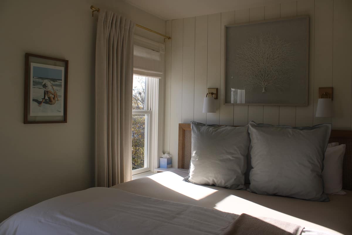 A bedroom with a large bed, a window with curtains, a wall-mounted lamp, and a framed picture.