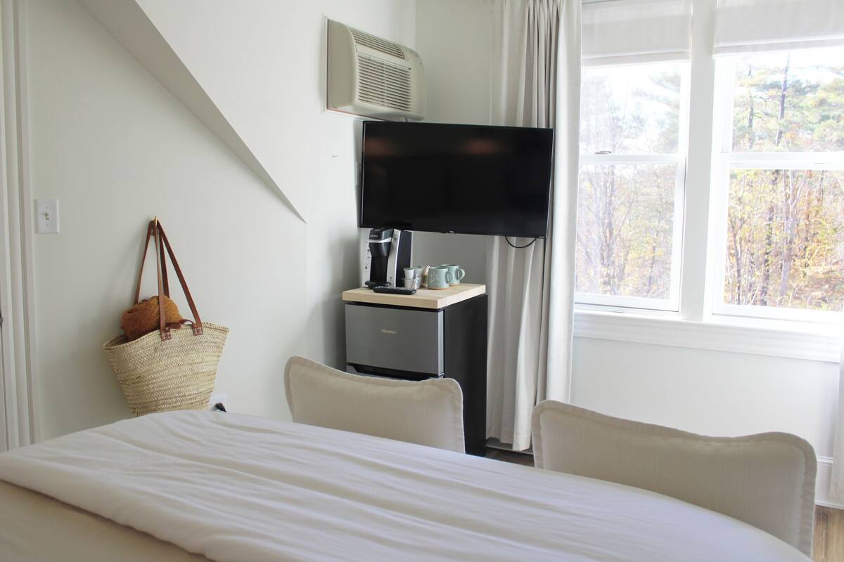 A hotel room with a bed, a desk with a television, a coffee maker, and a window with a view of trees.