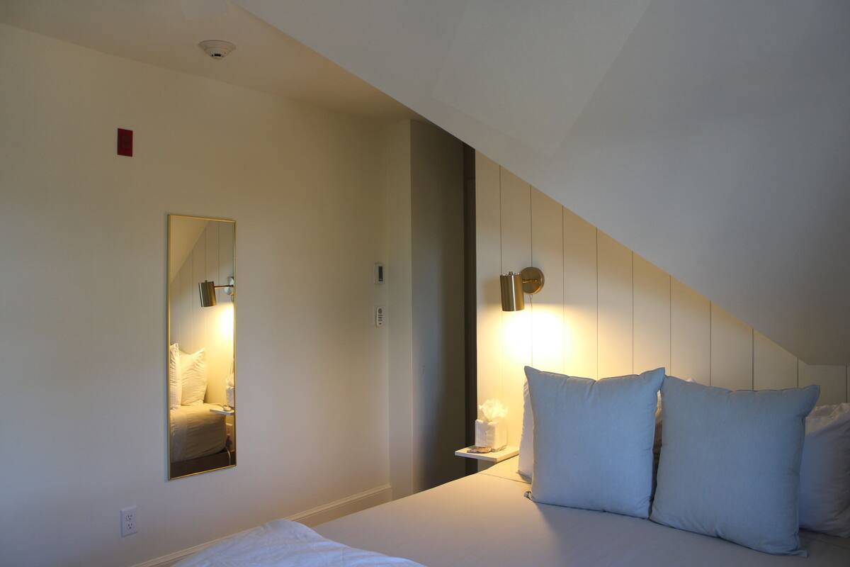 A hotel room with a bed, a bedside table, a nightstand, a mirror, and a wall-mounted light fixture.