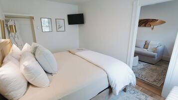 A bedroom with a large bed, a television on the wall, and a view into an adjacent living area.
