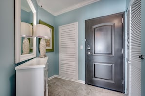 A hallway with a white console table, a mirror, a door with a handle, and a wall-mounted light fixture. - One Ocean Place 1209 - Garden City Luxury Oceanfront Condo (Garden City Beach)