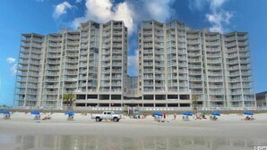 Et stort, mangeetagers hotel med en symmetrisk facade, talrige balkoner og beliggenhed ved stranden med parasoller og strandstole.