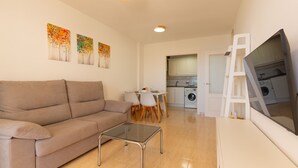Un salon comprenant un canapé, une table basse en verre et un coin repas avec des chaises et une table. On aperçoit une cuisine à l’arrière-plan, avec un lave-linge.