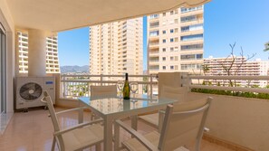A balcony with a glass table, chairs, a bottle, and glasses, overlooking a cityscape with high-rise buildings. - 6B Apollo XII sea view (Calp)