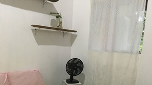 A bedroom with a bed, a fan, a window with a sheer curtain, and a shelf with pictures. - ESPAÇO RUSTIK  (Santa Cruz Cabrália)