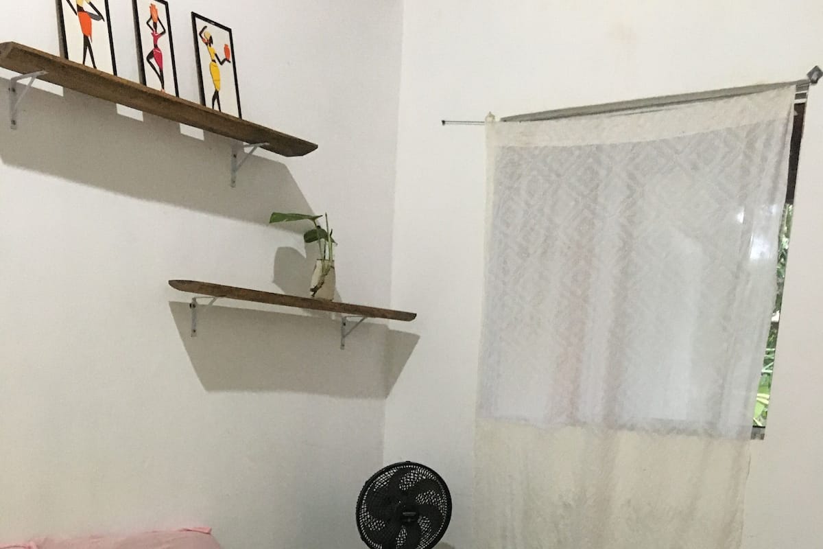 Una camera da letto con un letto, una ventola, una finestra con tenda leggera e uno scaffale con fotografie.