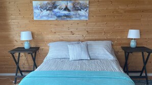 A bedroom with a wooden wall, a painting of swans, a bed with striped bedding, and two bedside tables with lamps. - Ocean view! Relax at this peaceful getaway only steps from the beach! (Grand Beach)