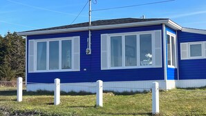 A single-story blue house with white shutters, a visible satellite dish, and a fenced yard. - Ocean view! Relax at this peaceful getaway only steps from the beach! (Grand Beach)
