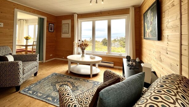 A cozy room with wooden walls, a round table with a vase, patterned sofas, and a view through large windows. - Ocean view! Relax at this peaceful getaway only steps from the beach! (Grand Beach)