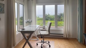 A room with a large window, a table with a vase of flowers, a white chair, and a wooden floor. - Ocean view! Relax at this peaceful getaway only steps from the beach! (Grand Beach)