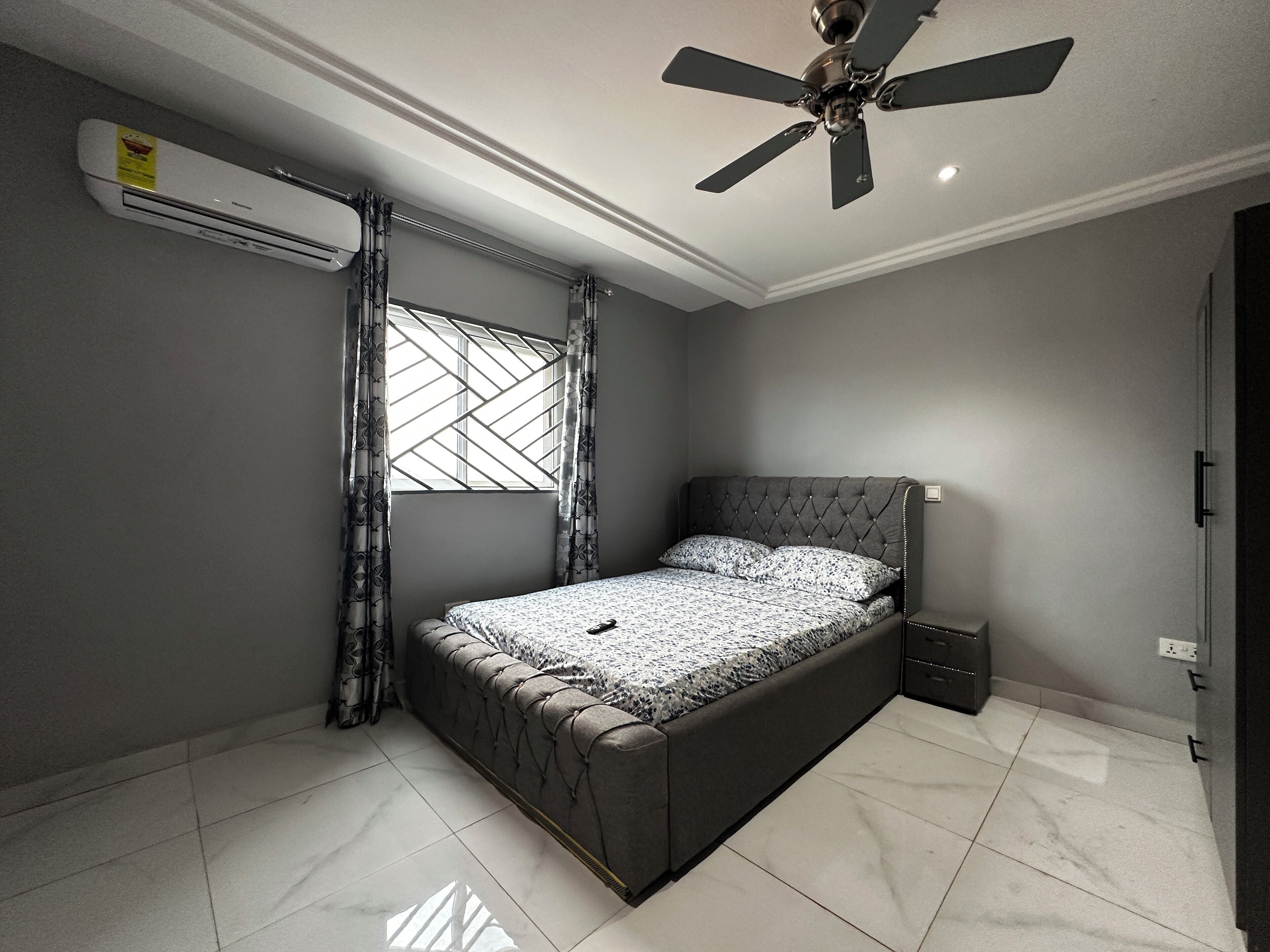 A bedroom with a bed, a ceiling fan, an air conditioner, and a window with curtains.