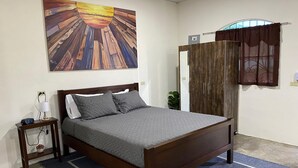 A bedroom with a wooden bed, a nightstand with a lamp, a wall-mounted painting, and a window with curtains. - Tres Cielos Cabanas (Playa Las Hojas)