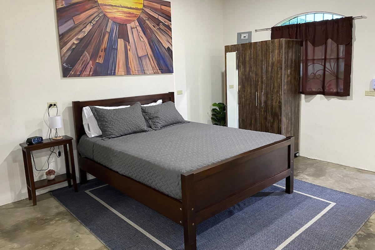 Een slaapkamer met een houten bed, een nachtkastje met een lamp, een wanddecoratie en een raam met gordijnen.
