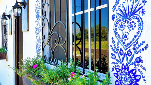 A building with a window featuring a decorative metal grille, a flower box with green plants and pink flowers, and blue floral patterns on the wall. - Tres Cielos Cabanas (Playa Las Hojas)