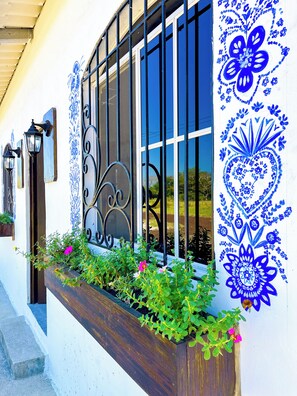 Un bâtiment doté d’une fenêtre ornée d’une grille métallique décorative, d’une jardinière contenant des plantes vertes et des fleurs roses, et de motifs floraux bleus sur le mur.