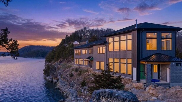 A modern two-story house by a lake with large windows and a stone exterior.
