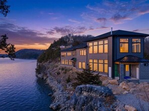 Une maison moderne à deux étages, située au bord d’un lac, dotée de grandes fenêtres et d’une façade en pierre.