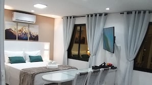 Une chambre d’hôtel compacte comprenant un lit, une table à manger avec des chaises, un petit canapé, un téléviseur fixé au mur et une fenêtre avec des rideaux.