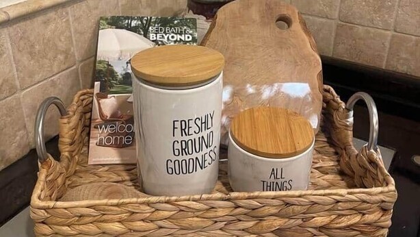 Un rincón de cocina con una bandeja tejida que sostiene un tarro blanco con la etiqueta 'FRESHLY GROUND GOODNESS', una tabla de cortar de madera, un tarro de vidrio y un libro titulado 'WELCOME HOME'.
