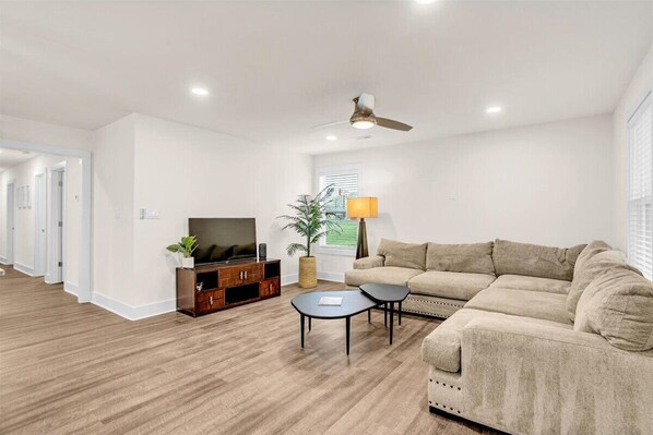 Una sala de estar moderna con un sofá beige, un televisor, una mesa de centro y un ventilador de techo.