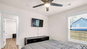 Un dormitorio con cama, ventilador de techo, televisor montado en la pared y ventana con persianas.