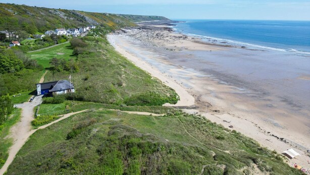 A coastal landscape with a sandy beach, a path leading to a building, and houses in the background.