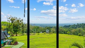 A view from a window overlooking a lush green landscape with a small white building in the distance. - Charming Jungle Bungalow - Coco Loco, La Fortuna. (La Fortuna)