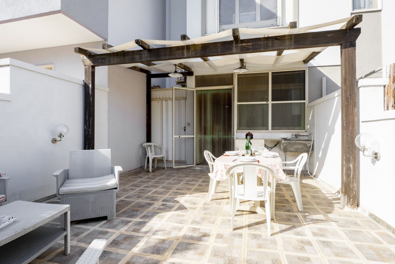 Une terrasse couverte avec une table et des chaises, un fauteuil en osier et un mur blanc avec une fenêtre.