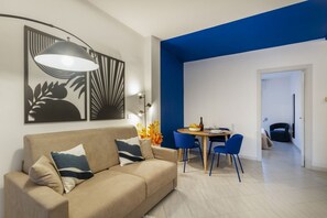 A modern living room with a beige sofa, blue chairs, a dining table, and abstract wall art.