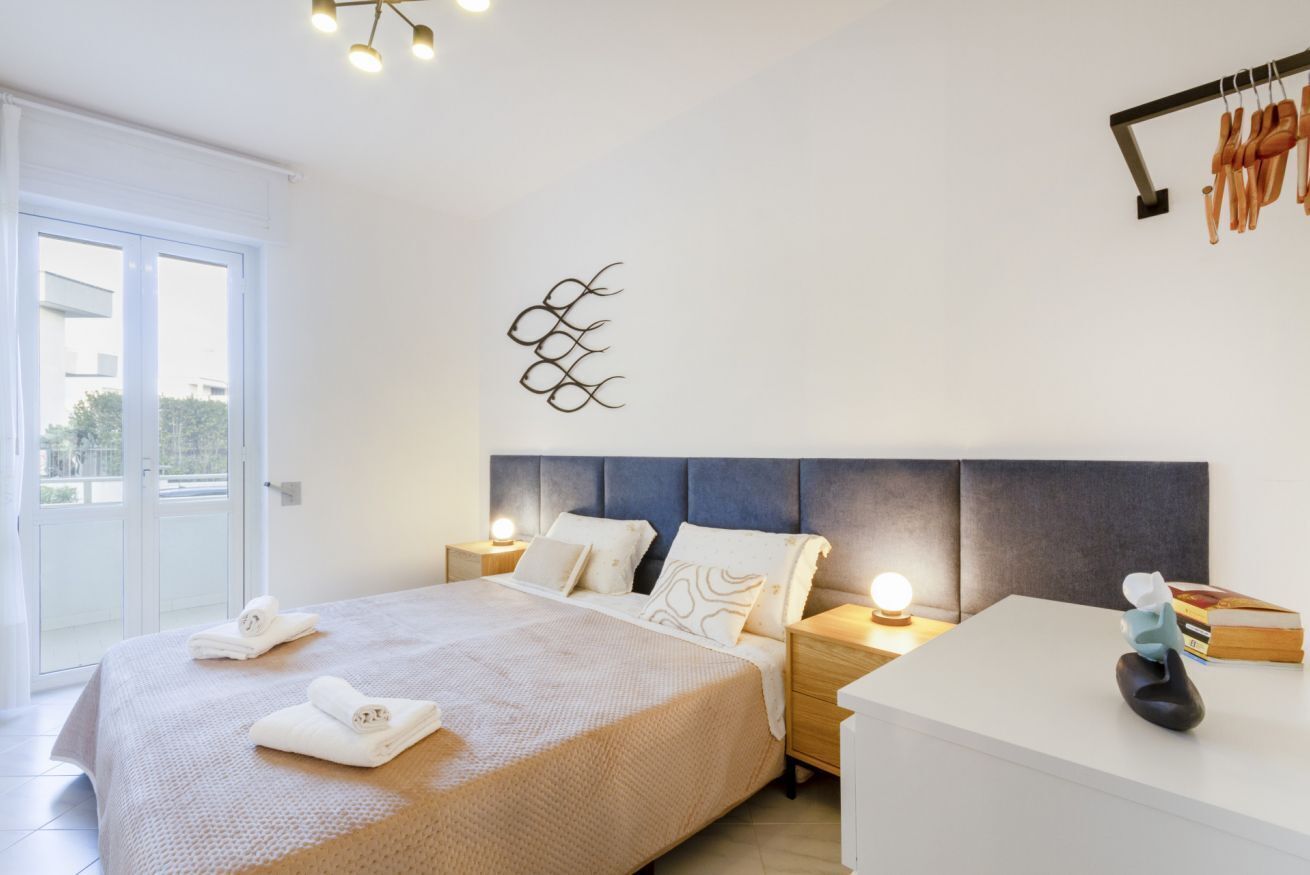 Un dormitorio con cama, mesitas de noche, una obra de arte en la pared y vistas a través de un ventanal corredera.